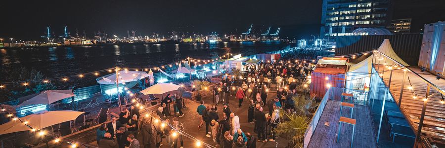 Abendliches Firmenevent am Platz am Fischereihafen mit Lichterketten und Hafenpanorama aus der Luft
